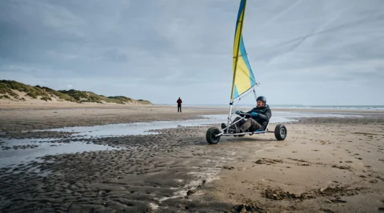Un débutant pilote un char à voile sur la plage plate du Cotentin à marée basse, sous un ciel nuageux normand.
