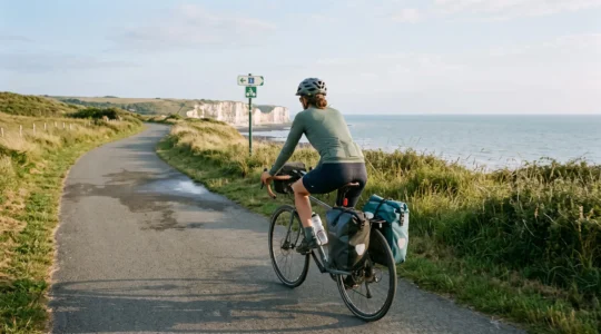 Un cycliste chargé de sacoches roule sur une voie verte côtière de la Vélomaritime en Normandie, falaises et mer visibles au loin.