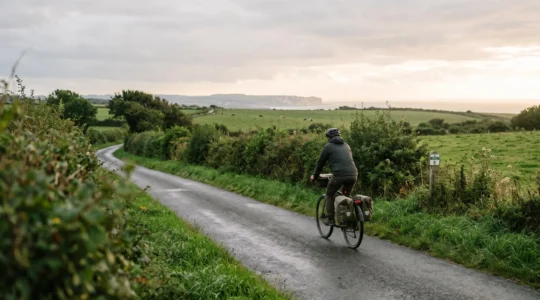 Un cycliste voyageur avec des sacoches roule sur une petite route de campagne normande bordée de haies, entre prairies et horizon côtier.