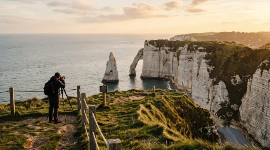 Un photographe photographie l'arche de la Porte d'Aval et l'Aiguille d'Étretat depuis le sommet d'une falaise de craie, derrière une barrière de sécurité, à la lumière dorée du soir.